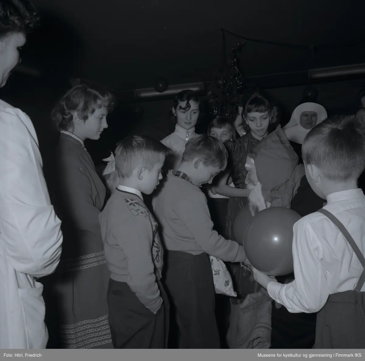 Den katolske menigheten i Hammerfest feirer juletrefest i 1957. En gutt har fått en gave fra julenissen som snakker til ham mens han slår ned blikket og ler. Andre barn og voksne står rundt dem og følger med.  
I bakgrunnen ser man juletreet og luftballonger.