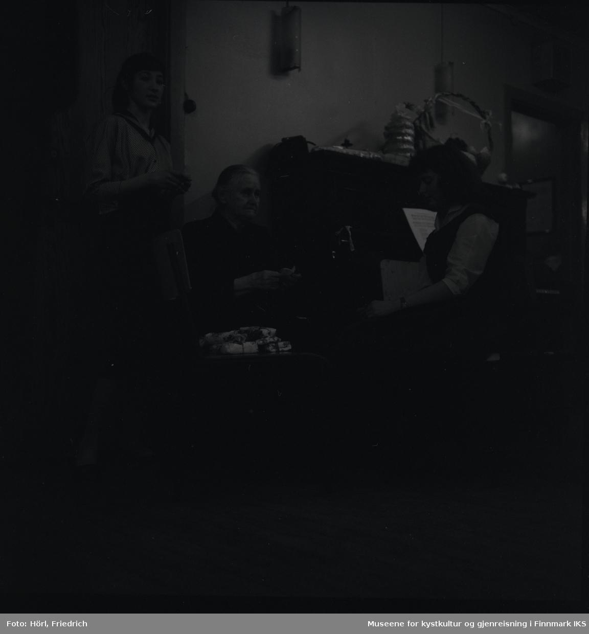 Tre kvinner er på juletrefesten i den katolske menigheten i Hammerfest i 1957. En av dem står, mens de andre to sitter. Mellom seg har de plassert en stol med gavepakker på. Ved siden av dem står en piano. På pianoet ligger det flere pakker, men også en kransekake og en kurv som er pyntet med sløyfer. Den eldre kvinnen i midten er Rønnaug Ottem Antonsen.