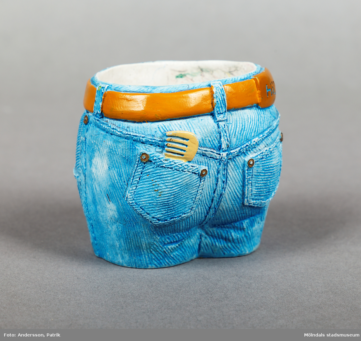 Pennställ i porslin, utformat som linningen på ett par jeans med ett bälte i midjan. Jeansen är ljusblå och bältet är ljusbrunt. Framtill har byxorna två fickor och en gylf. Baktill finns också två fickor. Ur en av bakfickorna sticker det upp en kam.