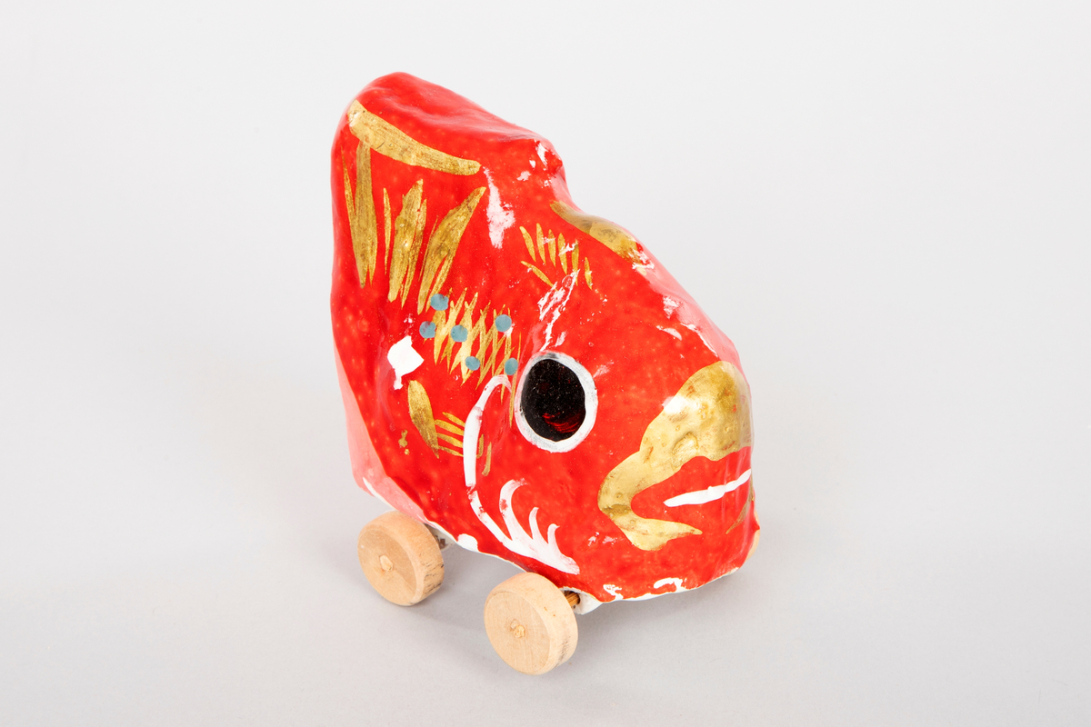 Fisk på hjul i pappmasjé og gips. Figuren er malt blank rød med store svarte øyne. Fisken har gullfarget munn, rygg og finne, noen blå prikker på kroppen og er matt oransje på undersiden. Hjulene er laget i tre.