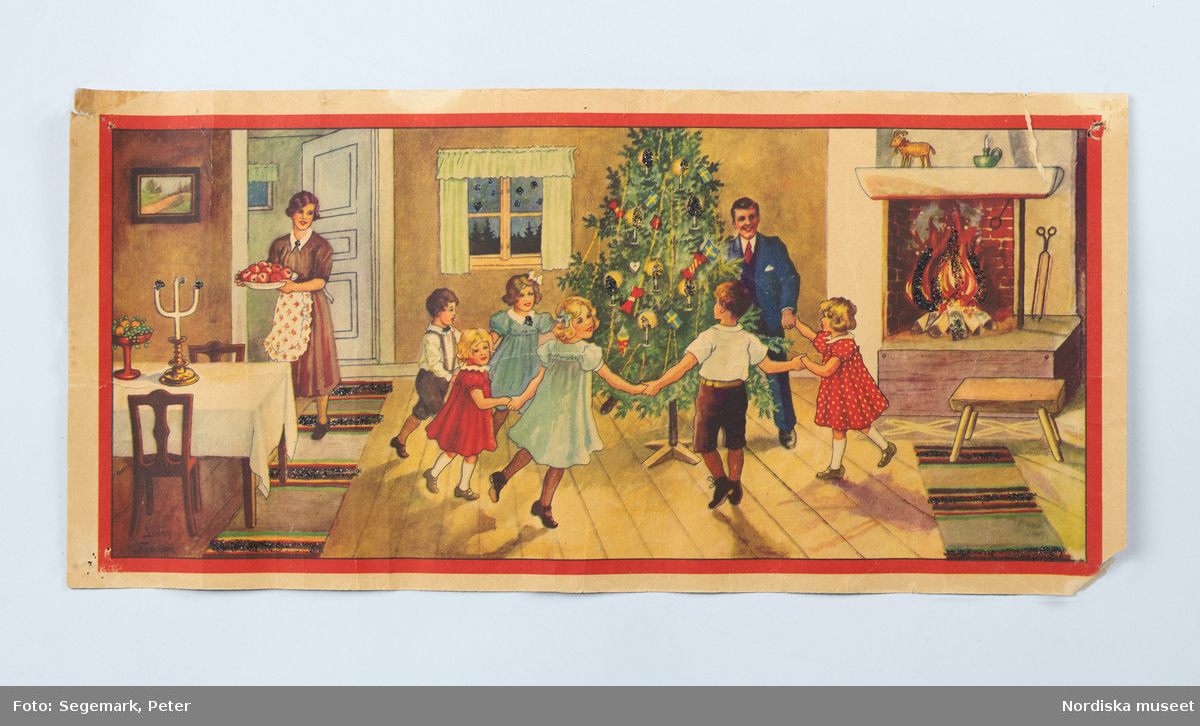 Plansch julfirande, utförd av Jenny Nyström. En man och några barn dansar kring julgranen. En kvinna bär ett fat med röda äpplen. I den öppna spisen är en brasa tänd.