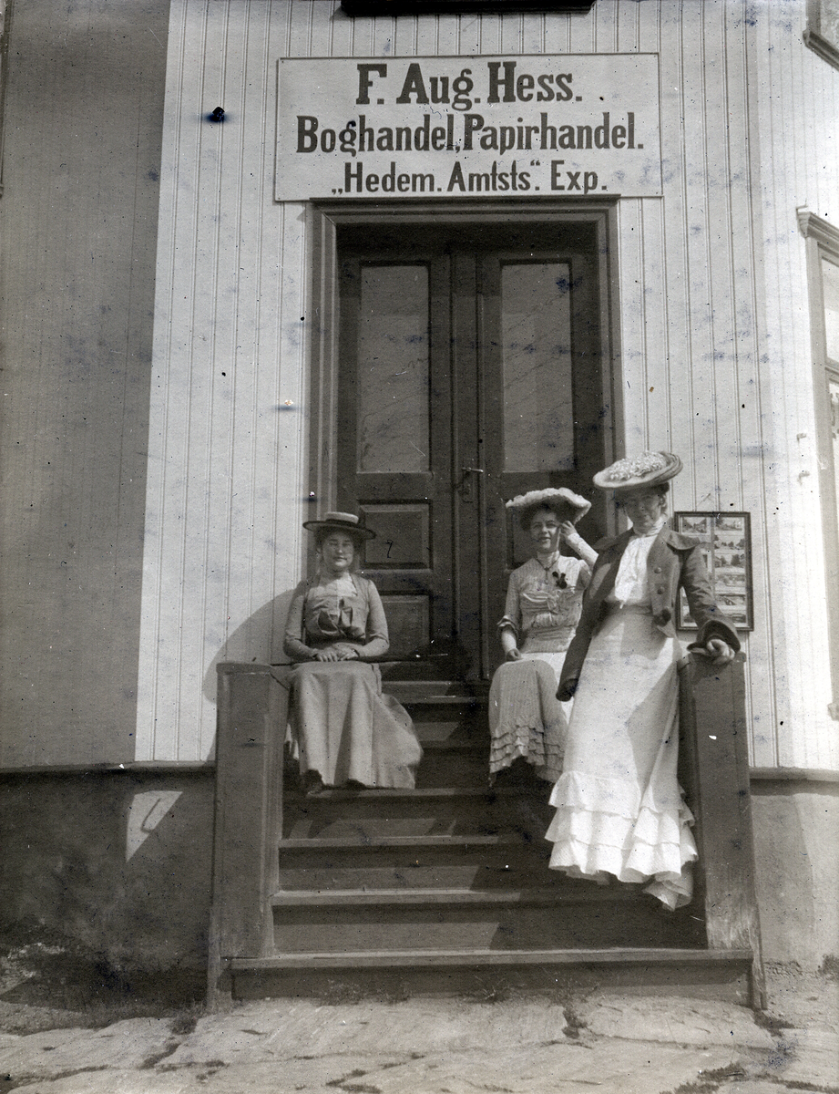 Tre kvinner står på trappa til F. Aug. Hess. Boghandel, Papirhandel "Hedem. Amtsts." Exp. Kongsvinger.
Sommerdag 1903.
Bilde er fra fotoalbum GM.036887.