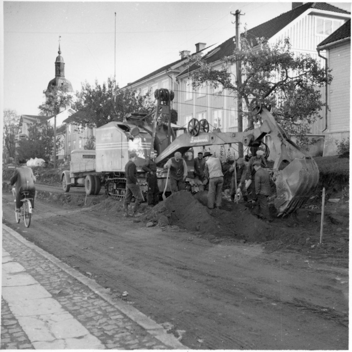 Åtta män vid en grävmaskin i arbete. En lastbil står bortom grävmaskinen. Södra delen av Brahegatan i Gränna breddas. En kvinna på cykel åker förbi norrut.