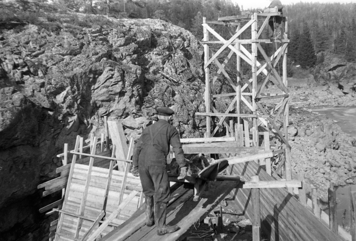 Fra bygginga av en ny dam ved Sølvstufossen, øverst i Ågårdselva i Tune i Østfold, som er en del av Nedre Glommas vestre løp. Anleggsarbeidet startet vinteren 1936, i en periode da det knapt rant vann i elveløpet. Dette fotografiet er tatt på tvers av vassdraget, fra nord mot sør, I forgrunnen ser vi en plattform med en anleggsarbeider som var i ferd med å tippe betong fra ei trillebår og ned i ei av forskalingene. Sentralt i elveløpet sto det et tretårn, antakelig et heisetårn, med en mann i toppen. Ågårdselva er et cirka fem og en halv kilometer langt vassdrag som renner fra den nordvestre enden av Isnesfjorden (Vestvannet) i Nedre Glommas vestre løp, sørvestover gjennem berglendt terreng mot innsjøen Visterflo. Høydeforskjellen mellom Isnesfjorden og Visterflo er på bortimot 25 meter. Ågårdselva har tre fossefall, det øverste her ved Sølvstu, deretter ved Valbrekke og nederst ved Solli. Behovet for den dambygginga vi ser på dette fotografiet var forårsaket av selskapene Borregaards og Hafslunds inngrep i Glommas østre løp, hovedløpet, som i lavvannsperioder gjorde det svært vanskelig å få tømmer som skulle til bedrifter nedenfor Sarpsfossen via Mingevannet, Isnesfjorden og tømmertunnelen til Eidet gjennom det trange sundet ved Trøsken. Dette forsøkte man å løse ved å heve vannspeilet i Isnesfjorden med en dam som skulle plasseres 10-15 meter ovenfor den eksisterende dammen ved Sølvstufossen. Se mer informasjon under fanen «Opplysninger».