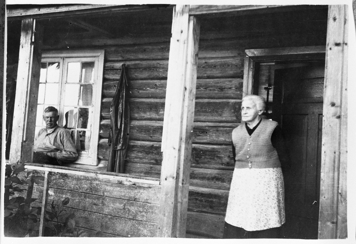 Eldre mann og kvinne avbildet foran inngangen til tømmerhus. Ingen informasjon foreligger.