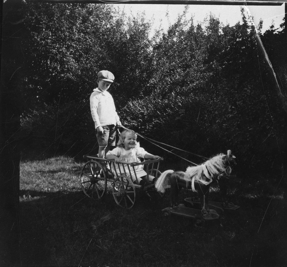 Foto av søsknene Nils Fredrik og Cecilie Aall utenfor lekende i vogn med lekehester, august 1916