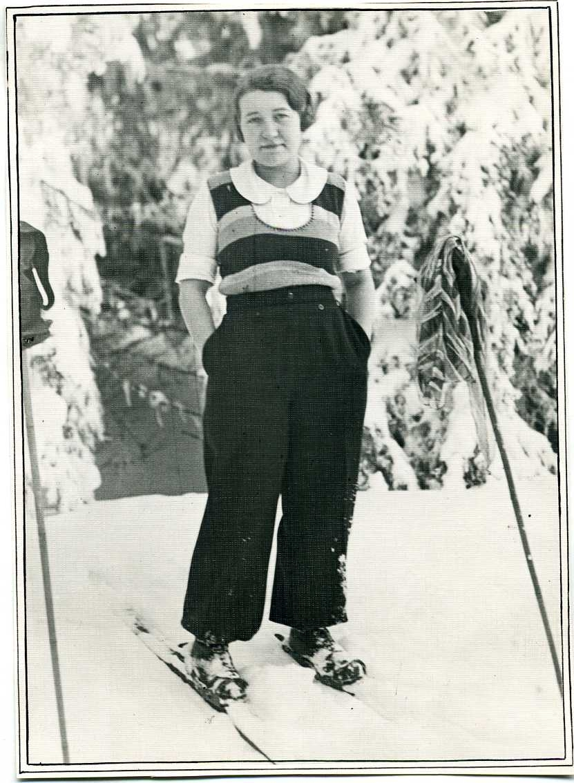 Ett vinterlandskap. En ung kvinna som står på skidor, med händerna i byxfickorna. Hon bär vida mörka byxor och en randig väst ovanpå en kortärmad tröja.