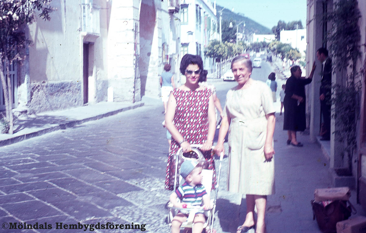 Tomas (?) mamma och mormor gåendes på en gata. Pojken sitter i en sulky. Fotografiet ser ut att vara taget utomlands. Även årtalet är okänt.