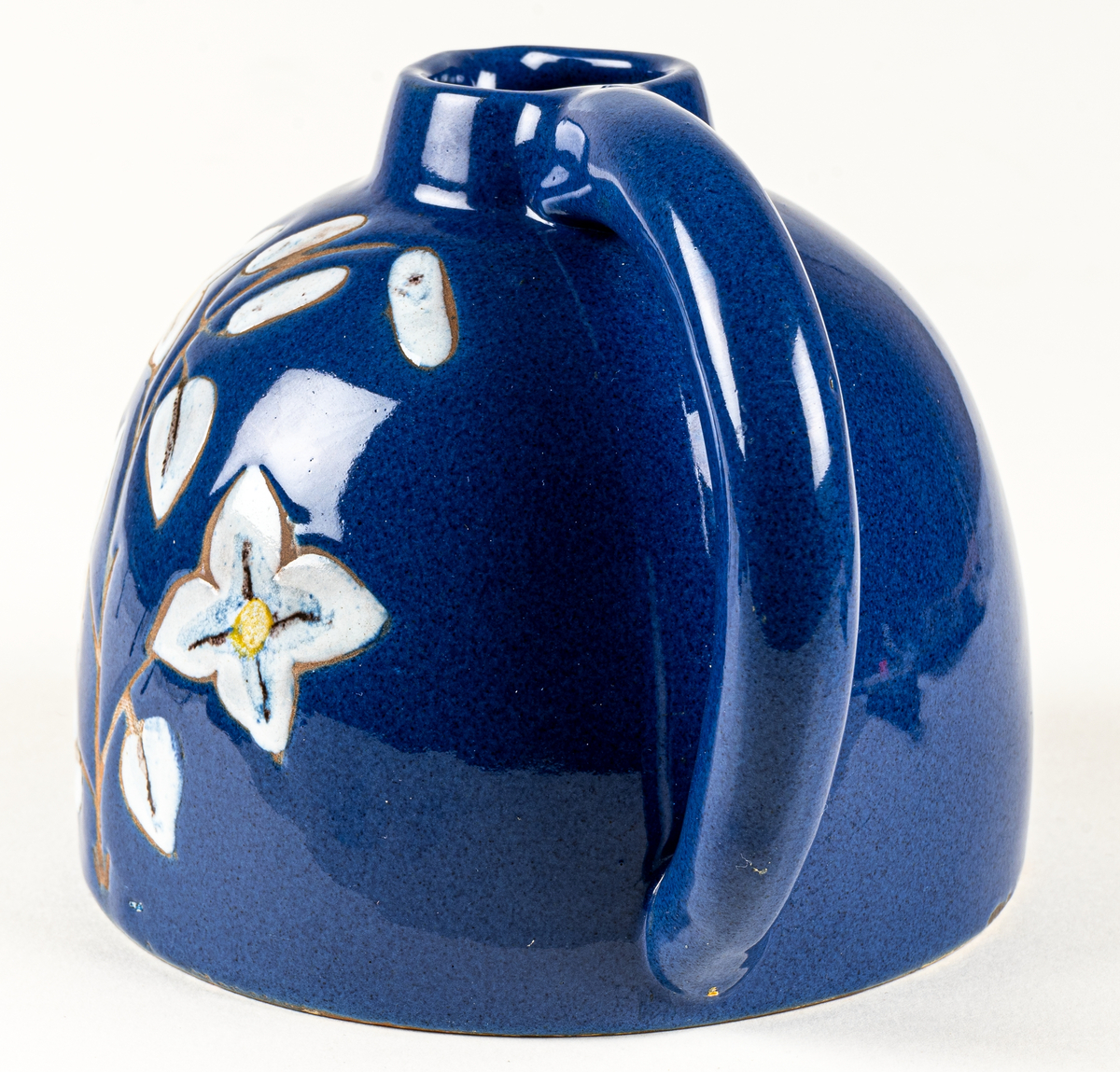 Kanna eller blomkrus i lergods, formgivet av skulptör Maggie Wibom vid hennes företag Stockholms Keramik AB, 1940-talet. Den blå glasyren fick namnet Wibomblå, kannan dekorerad med en kvist vita blommor där kvisten och konturen är oglaserad genom påläggning av vax innan glasyrbränning.