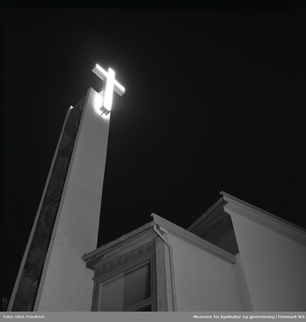 Det lyssatte korset ved klokketårnet til den katolske kirka St. Mikael i Hammerfest lyser i nattemørke. Bildet er tatt oppover fra gateplan i Strandgata.