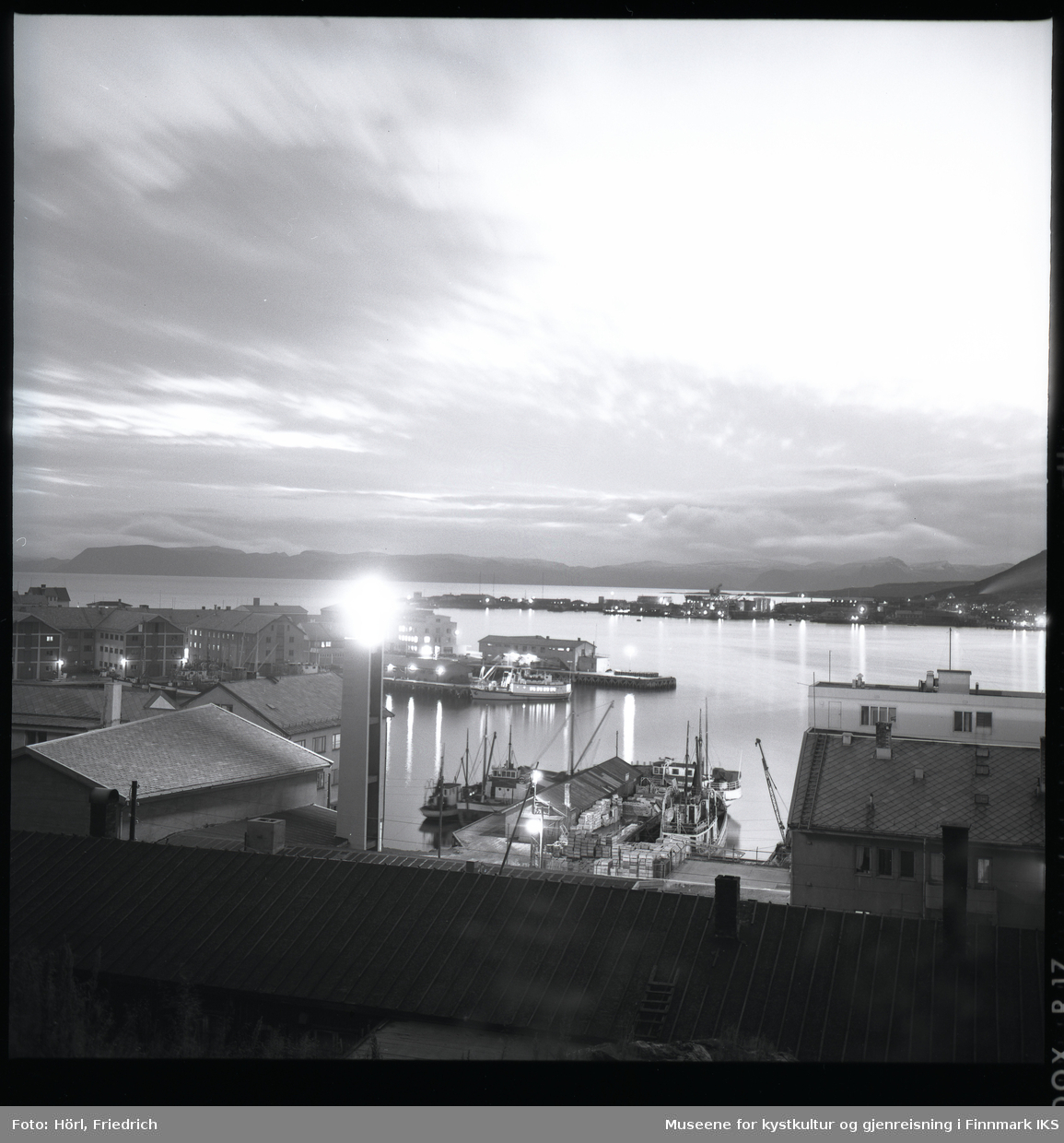 Det er skumringstime i Hammerfest og fotografen tar et bilde fra fjellsiden av Salen ut mot havna. I forgrunnen ser man et brakketak. En lys flekk på bildet er der det lyssatte korset ved klokketårnet til den nybygde katolske kirka St. Mikael er. Gatelysene er på. Ved kaia ligger det båter og på andre siden av bukta ser man bydelen Fuglenes. Lengst bak i bildet ser man silhuetten av Sørøya.
