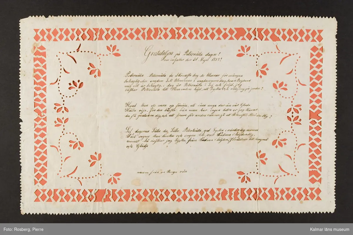KLM 20679. Gratulationsbrev, silhuettklipp. Av papper, färglagt i rött. Text, bl a: Gratulatjon på Petronälla dagen, den 31 Majus, 1858. Ornamenten urkippta.