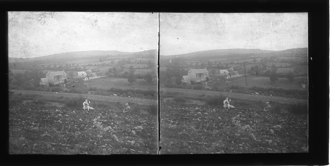 En kvinna, en man och hund sitter i en slänt - plöjd åkermark? Nedanför dem går en järnvägsräls och på andra sidan ligger en större gård, man anar en fylld luftballong?
Diapositiv, stereoskopisk bild.