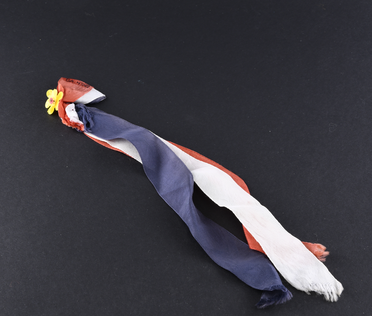 17. Mai sløyfe. Består av tre tekstilbånd i rødt, hvit og blått som er lagt dobbelt i øvrekant. Båndene er festet med nål som øverst har en gul blomst av plast. Nedre deler av båndene har skrå sårkanter.