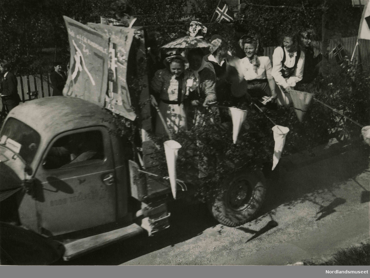 En lastebil pyntet med bjørkekvister. I lasterommet står det unge kvinner i pene klær. De holder noe som ligner på håver. Bilde tatt i Bodø under feiring av freden etter okkupasjonen 1945.