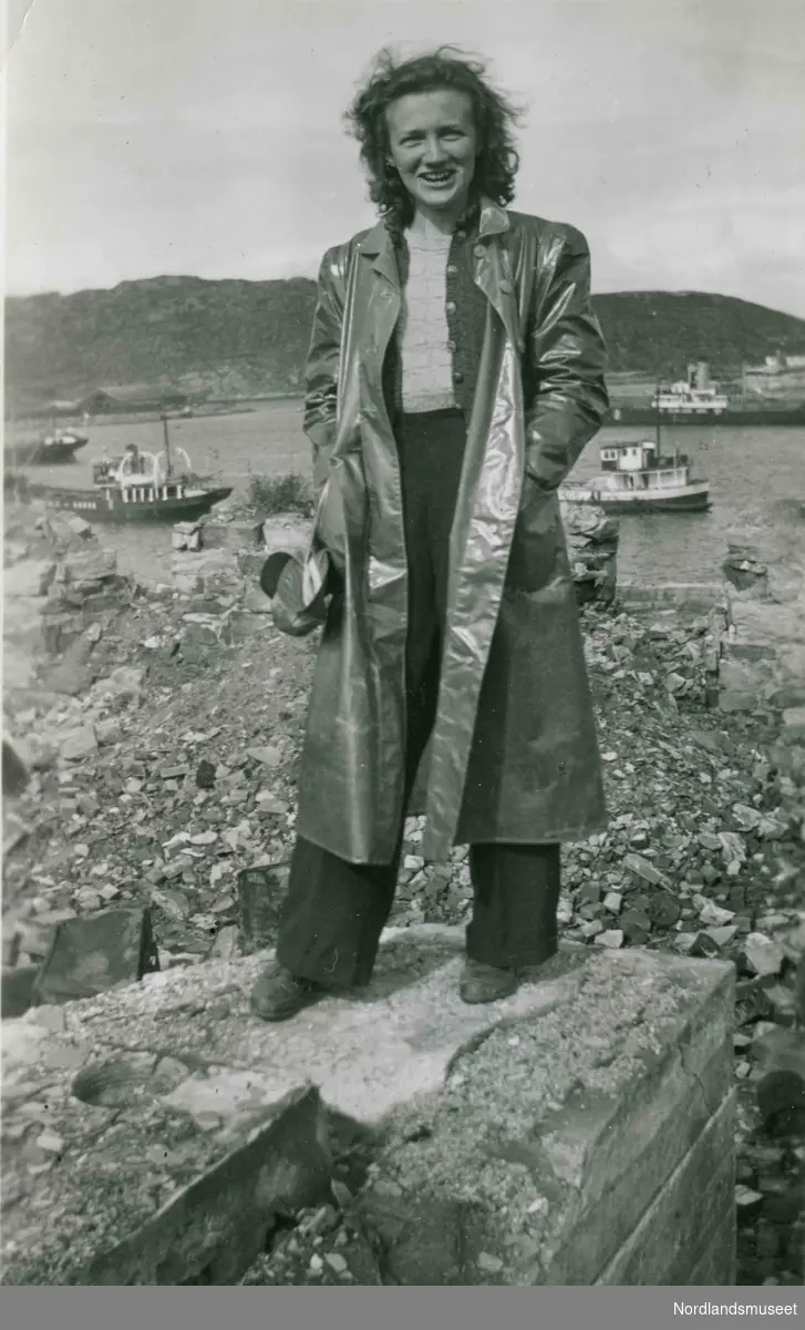 En ung kvinne i regnfrakk står på restene av en mur. I bakgrunnen båter. Bildetekst: "På gamle tomter. August 1942. Huset i Sjøgata 4 som ble bombet".