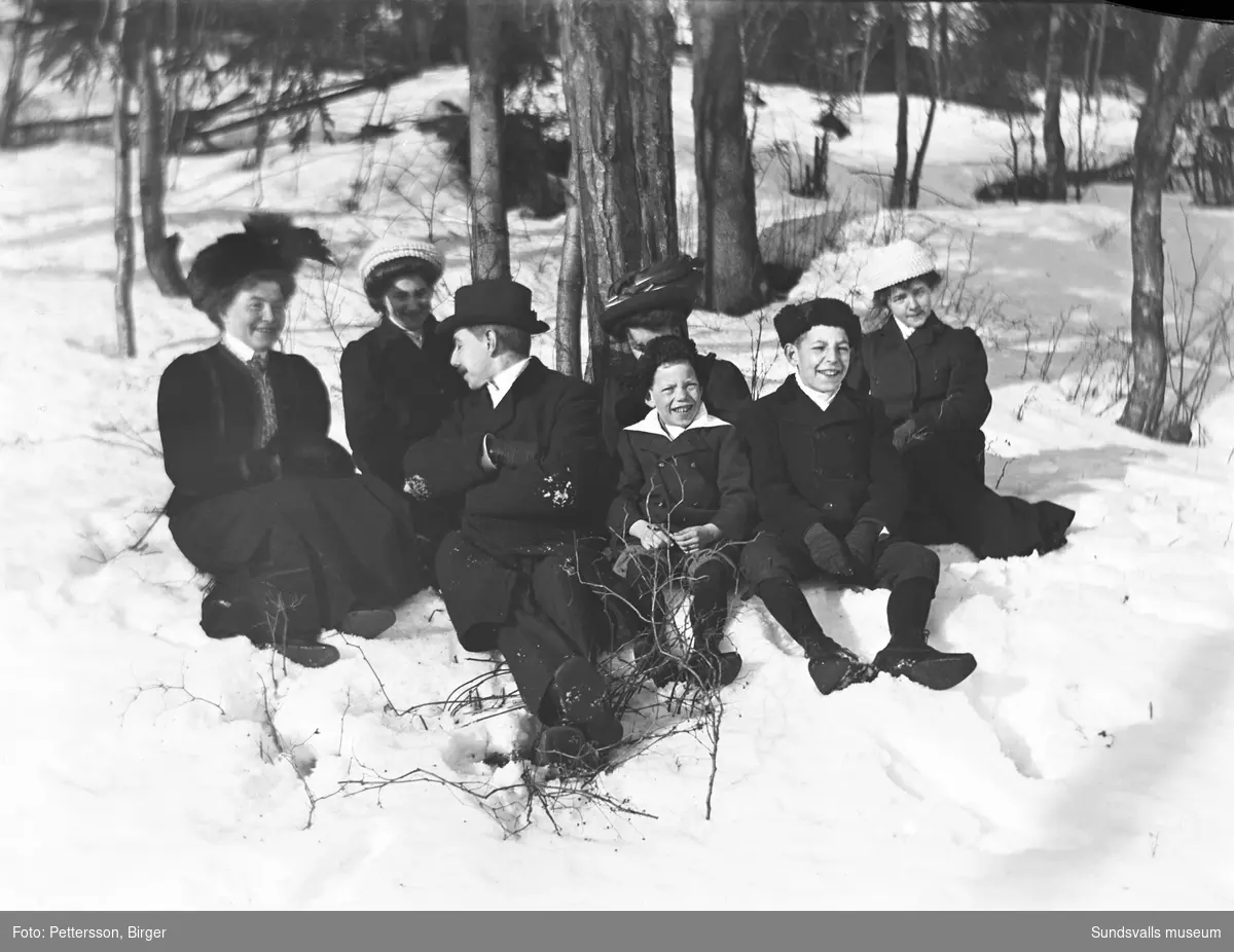 Gruppbild med fyra kvinnor, en man och två barn som sitter i en snödriva i skogskanten.