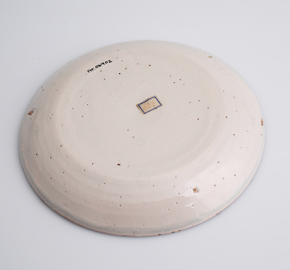 En flat tallerken av keramikk/porselen. Den har bly- og tinnglasur og er hvit på farge. Tallerkenen er rund med en relativt høy og skrånende kant. Den har ikke dekor utover glasuren. Tallerkenen er en av 11 identiske tallerkener som trolig er fra tidlig på 1700-tallet.