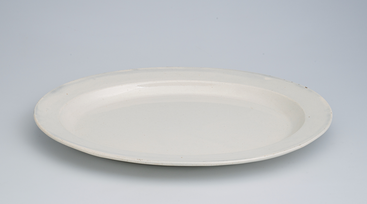 Et lite ovalt serveringsfat av glasert keramikk/porselen. Det er ensfarget hvitt (med hint av lyst blågrått i) på farge. Det er et enkelt fat uten særlig dekor. Det er stemplet på undersiden.