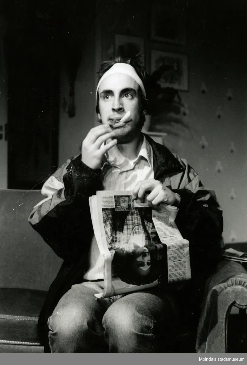 Nya Teaterhuset okänt årtal. Skådespelaren och komikern Per Andersson vid en repetition eller föreställning. Han sitter på en stol och har något på huvudet samt en cigarett i munnen. I knäet har han en tidning.