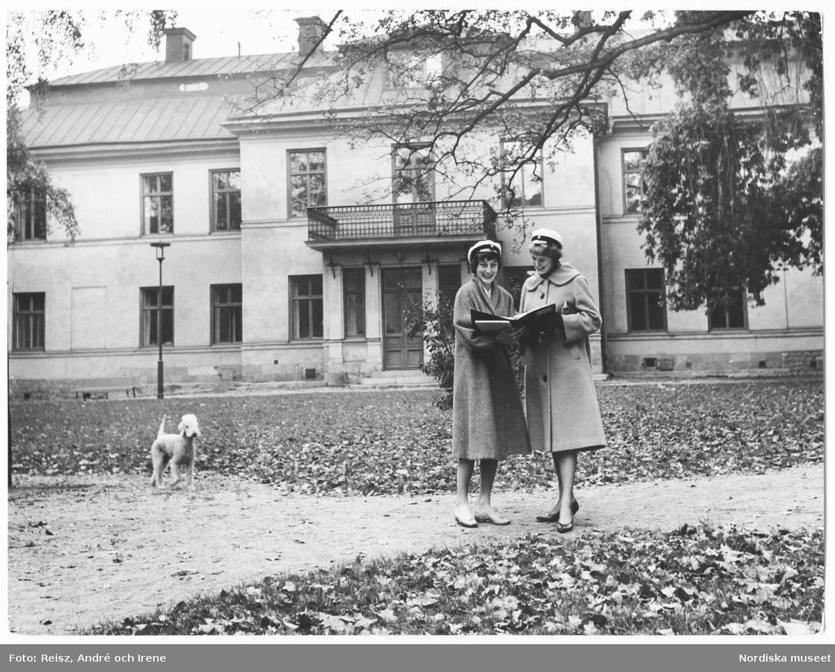 Stockholms universitet. Två unga kvinnor i studentmössor med en hund i sällskap framför en av byggnaderna på området.