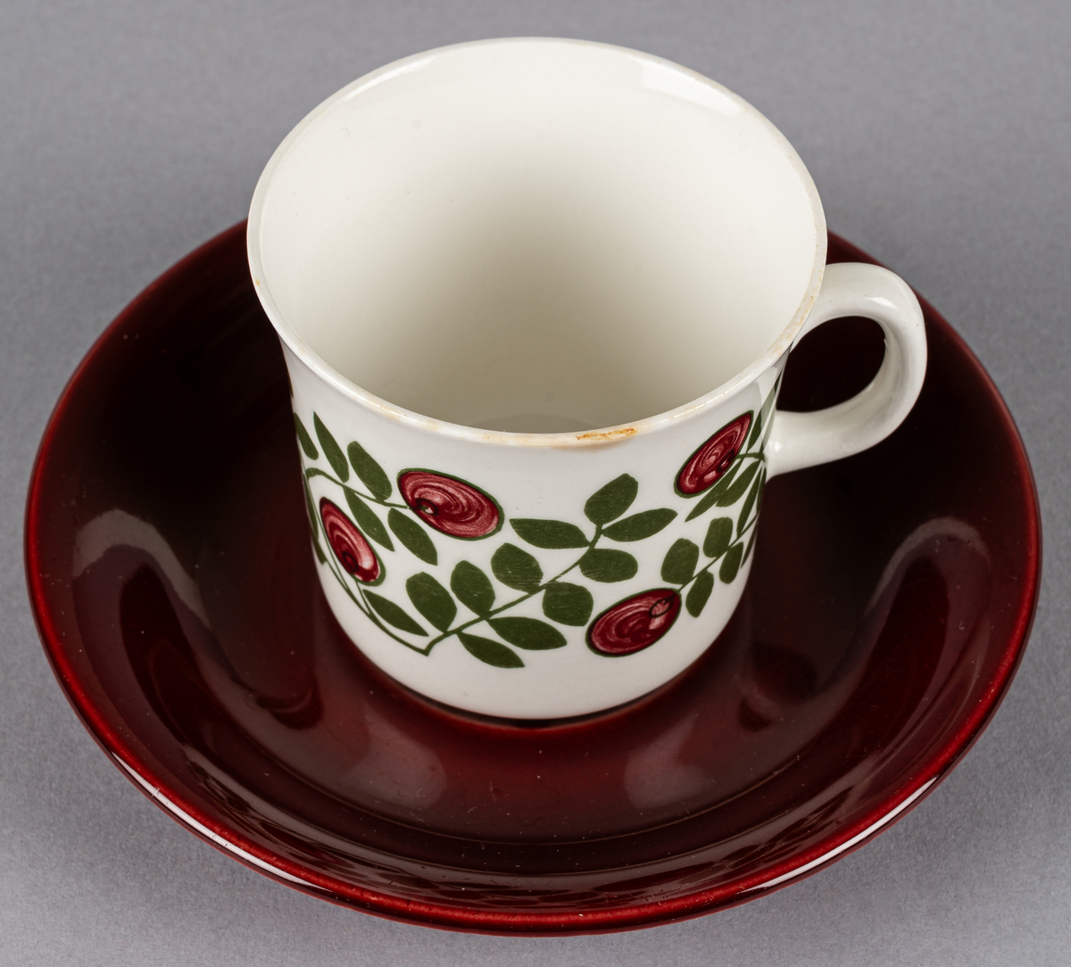 Kaffekopp med fat, i flintgods. Modell EF, dekor Klarbär. Vit glasyr med röda bär och grönt bladmönster i grönt, fatet mörkrött. Otydlig stämpel på fatets undersida. Tillverkad troligtvis på 1960-talet.