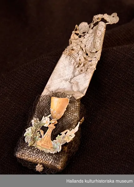 Av silverpapper med genombrutet mönster i kanten. Påklistrad tyllbit med stycke i form av en urna och blomsterkrans.