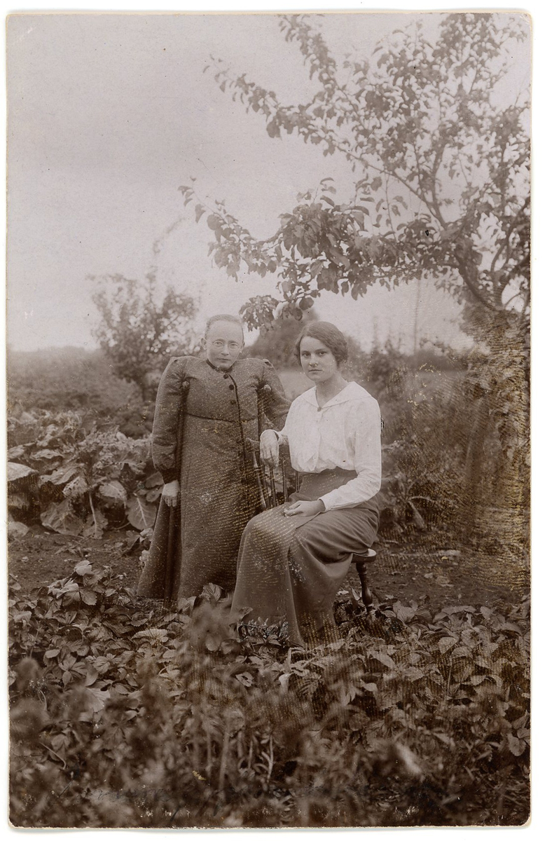Två kvinnor i en trädgård.
Den ena kvinnan sitter på en stol. Den andra kvinnan, Selma Ljungberg, står stödd av två käppar. Hon är rörelsehindrad, kort och saknar ett ben.