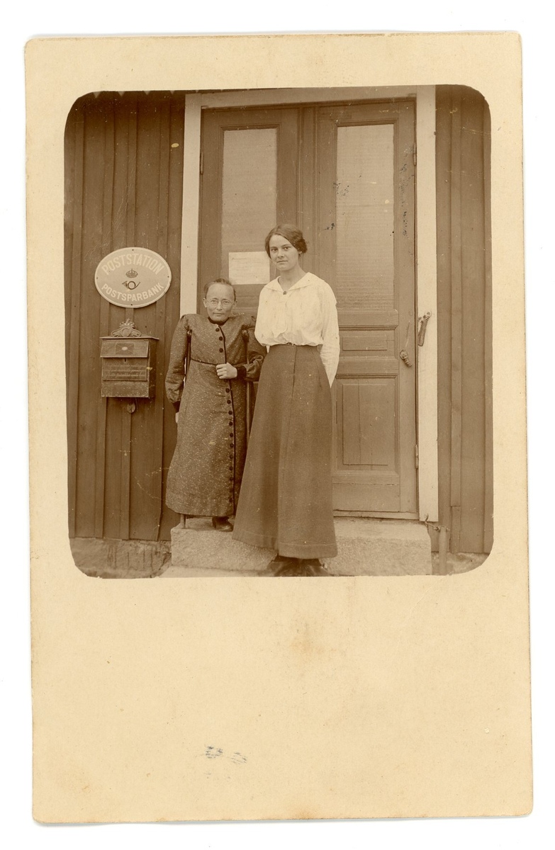 Två kvinnor stående utanför ett hus med poststationsskylt och brevlåda. Den ena kvinnan, Selma Ljungberg, är kort, ser ut att sakna en fot och stödjer sig på två käppar.