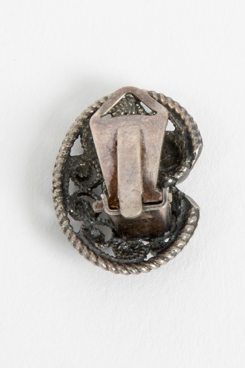 En av to øreklips i sølv. C-formet med filigransarbeid, tvinnetråd langs kantene og åtte små og to større sølvperler.
