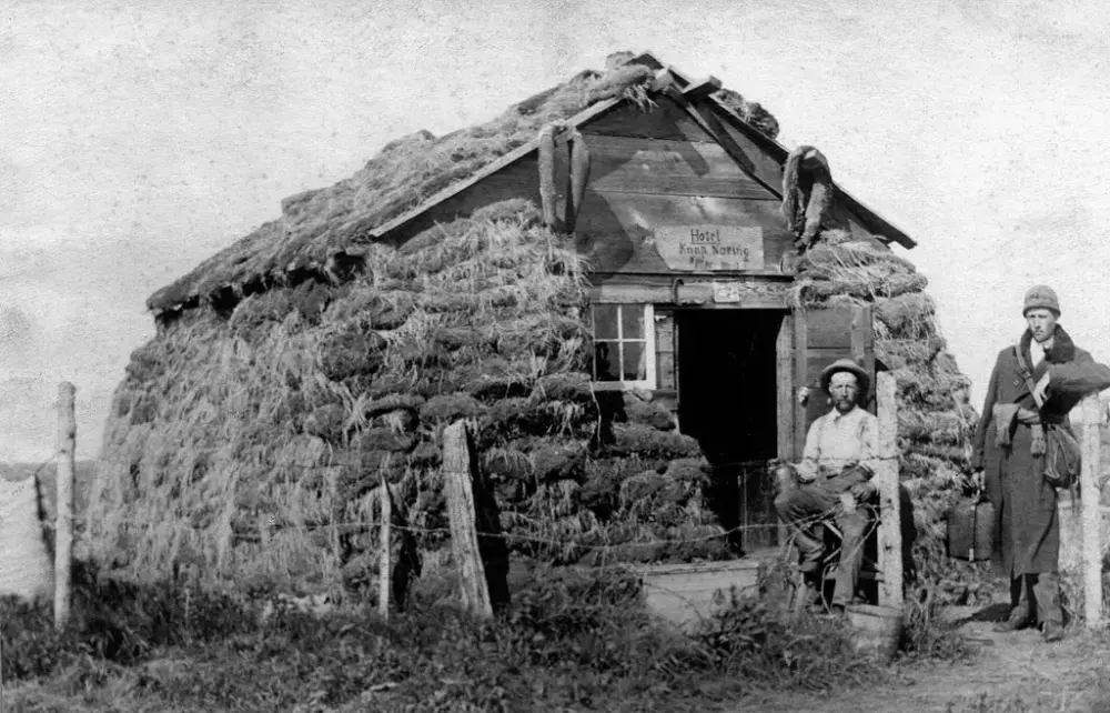 Historisk eksteriør av et hus bygget av torv. Deler av gavlveggen er i tre. Et lite vindu og en åpen dør i gavlveggen. En mann sitter i døra. En annen mann i frakk og med tatt og koffert står til høyre og lener seg på en portstolpe.