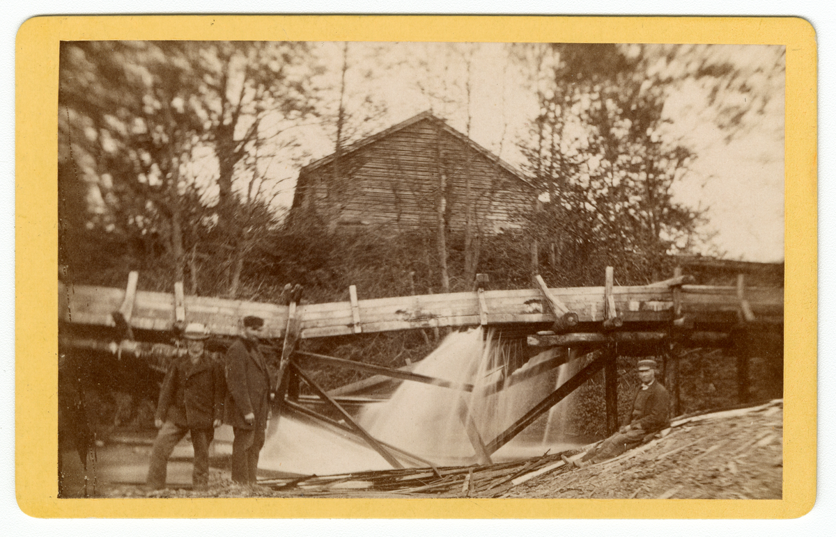 Foto av menn foran en "akvedukt" antagelig på Dal gård i Ullensaker 1880-tallet. Vannsystemet ser ut itl å leke