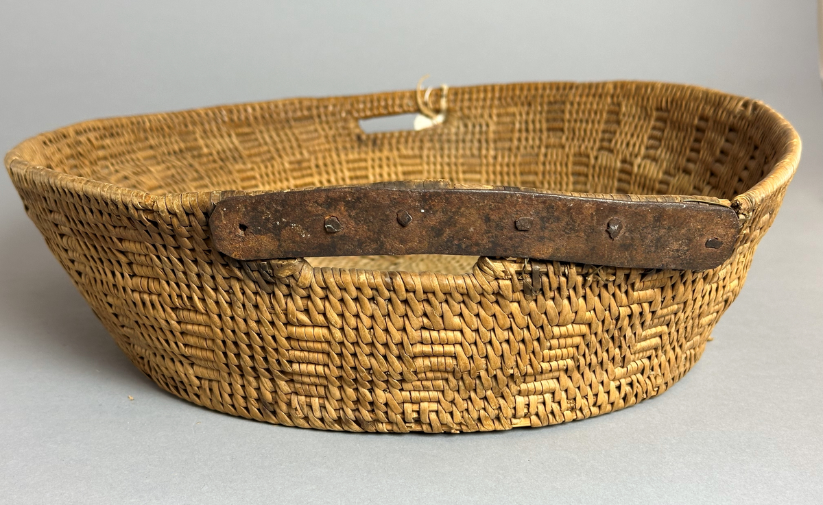 En oval såkorg, tätbunden av rot med mönster. Två handtag, varav det ena är lagat med en metallskena.