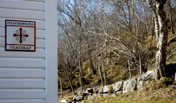 Skilt med teksten pilgrimsleia og vertskap med pilgrimslogoen på montert på hvit trevegg. Steinutgard i bakgrunnen.