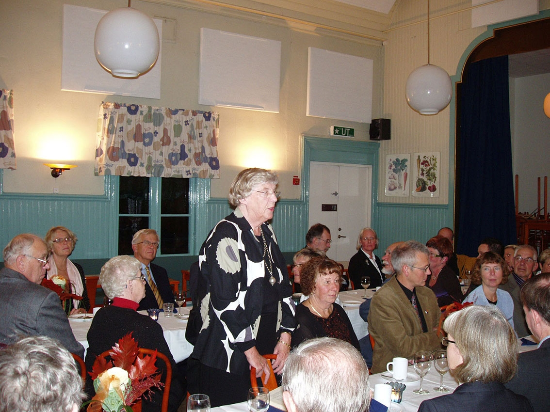 En kvinna står upp vid ett långbord och håller ett tal till sittande middagsgäster. Kvinnan är tidigare ordförande vid Stiftelsen Grenna Museum.