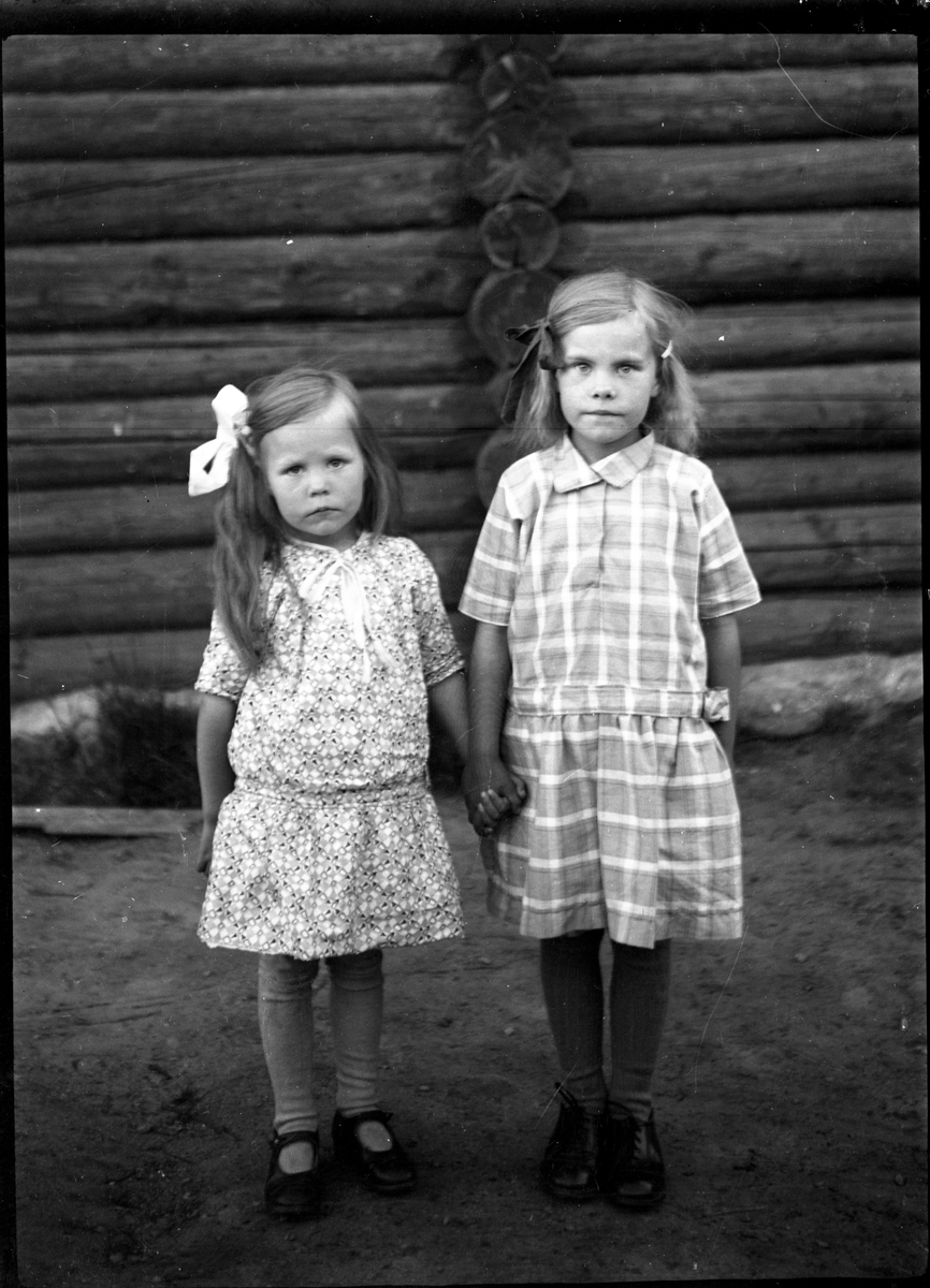 Portrett to jenter som holder hender foran tømmervegg. Antagelig Ole Romsdals barn 

Fotosamling etter fotograf og skogsarbeider Ole Romsdalen (f. 23.02.1893).