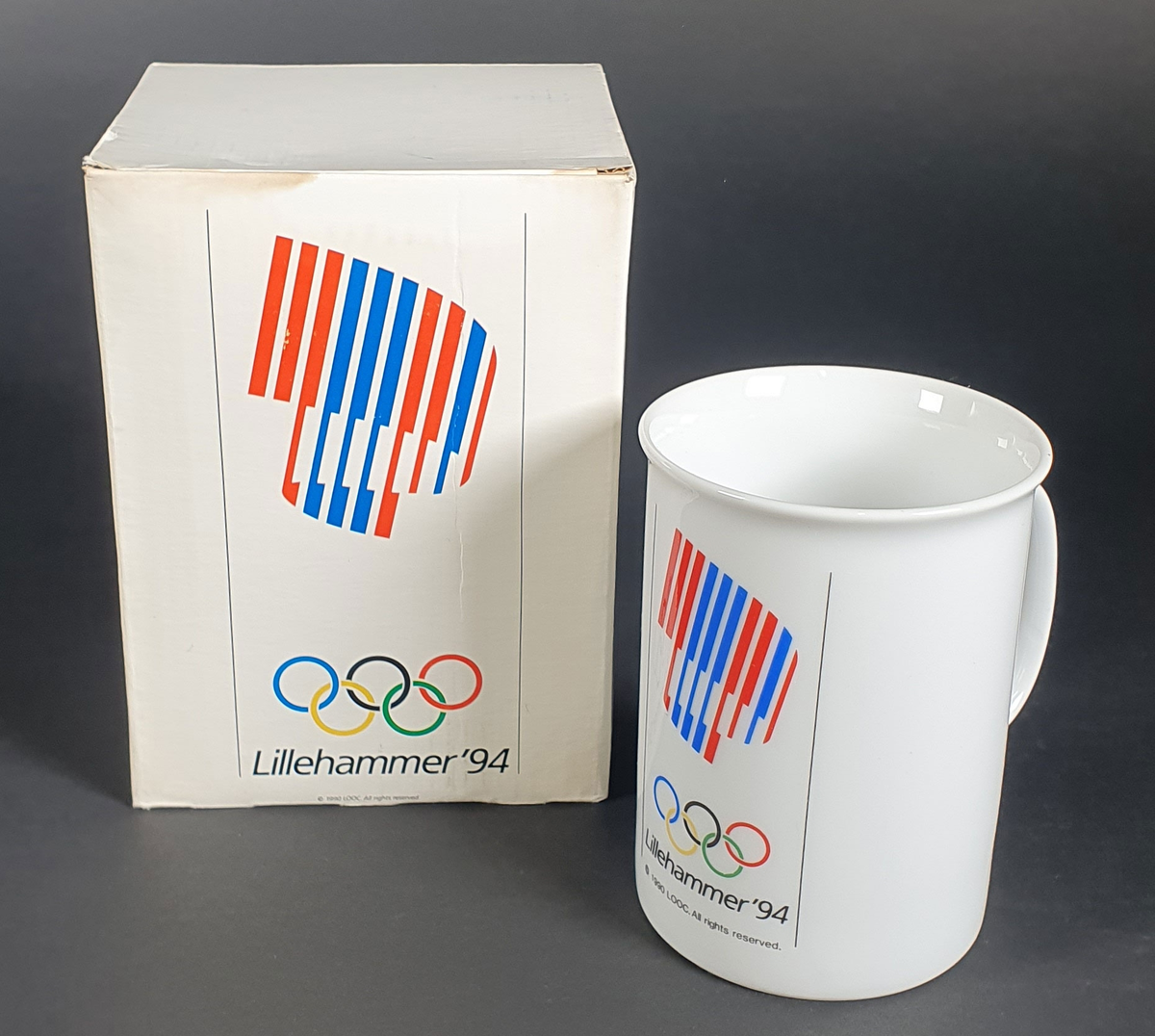 Hvitt kopp med hank, og med emblem for Lillehammer '94 i rødt, hvitt og blått. Koppen ligger i original emballasje.
