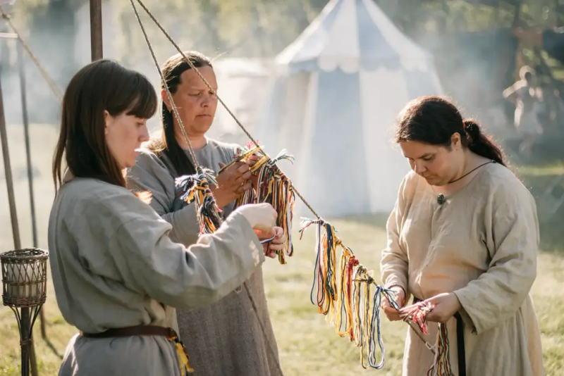 Tre kvinner i hvite middelalderlinkjoler som lager armbånd i gult, rødt, grønt, og blått.