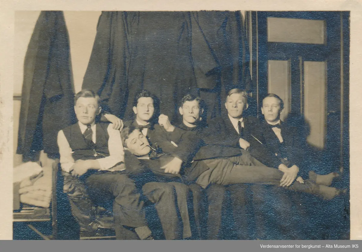 Et gruppebilde av 6 menn i en garderobe eller klasserom, det henger flere jakker på veggen og mennene sitter i en sofa under dem. En av mennene ligger i fanget til de andre med en pipe i munnen.  