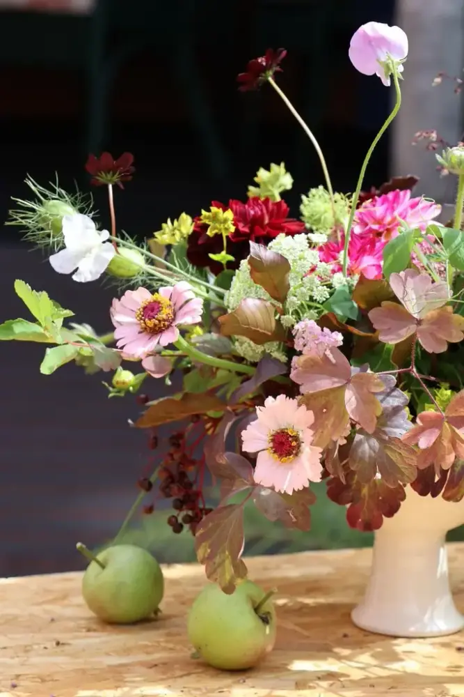 En blomsterbukett i vase og to epler på et bord. 