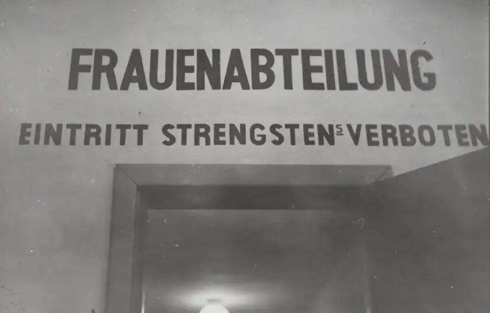 Fotografi svart/hvitt tatt inne i Grini fangeleir, inne i murbygningen ved inngangen til Kvinneavdelingen. Man ser en dør som er åpen og over døra står det "FRAUENABTEILUNG. EINTRITT STRENGSTENS VERBOTEN".