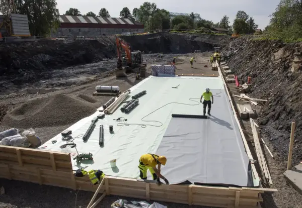 Firkantet, stor forskalingskasse hvor en oransje gravemaskin jevner ut grus mens flere arbeidere legger på markisolasjon før støping.