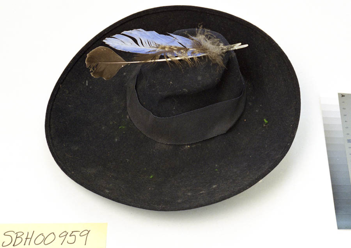 Form: Hatten er rund og har en bred brem. Bremmen er brettet oppover rundt halve hatten. På samme side henger det to bånd ned fra undersiden. Bredt hattebånd i samme materiale som disse båndene. På toppen er det sydd på tre fjær. (I den midlertidige katalogen står det at det hører til en hattenål også. Det er mulig at nålen som er nevnt ved nummer SBH.00956 er den som er ment her, i og med at den nålen lå løst. Men på denne hatten er det ikke spor etter nålen). På kanten av bremmen går en søm. Og langs kanten inni, rundt åpningen, er det sydd på et bredt bånd, og en filtrem som kan festes under haken.
