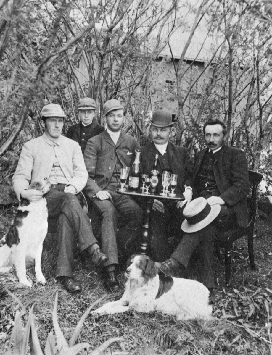Fem menn rundt bord utendørs med glass og flasker på bord. To hunder. Løken østre, Rudshøgda, Ringsaker.