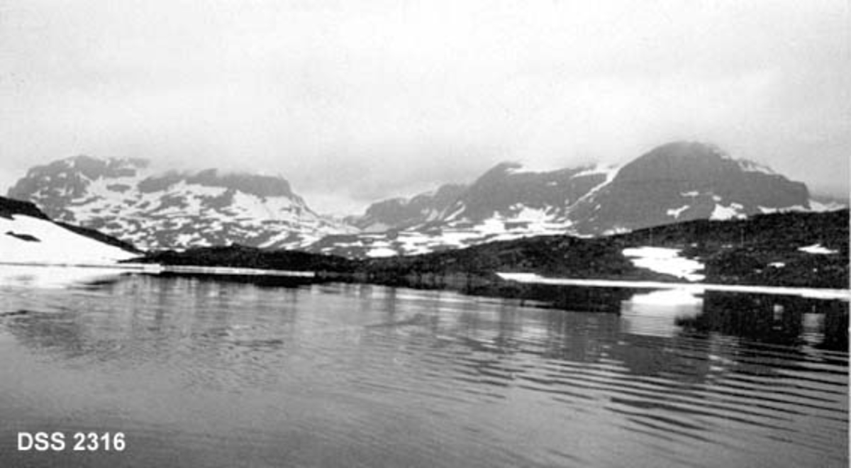 Utsikt fra Haukelivatnet mot Dyrskar.  Høgfjellslandskap med sjøflate i forgrunnen, bergrabber og snaufjell med snøstriper i bakgrunnen, tåke over toppene. 