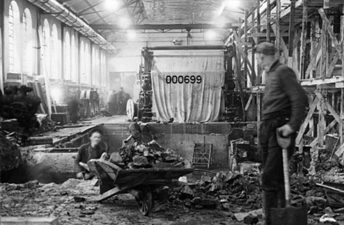 Interiør fra A/S Buskerud Papirfabrikk på Åssida i Drammen (den gang i Lier kommune).  I 1937 ble papirmaskinen ved denne fabrikken fullstendig ombygd, særlig for å kunne kjøre maskinen med større driftshastigheter og dermed oppnå større produksjonsvolumer.  Fotografiet viser den indre delen av maskinsalen, der hvor papirmaskinens tørkeparti hadde stått.  Da fotografiet ble tatt var denne delen av maskinen demontert, og arbeiderne arbeidet med å tømme maskingrava for bygningsavfall.  I forgrunnen står en arbeider med spade ved sida av ei fullastet tretrillebår i samtale med en mann som arbeider nede i maskingrava.  Sentralt i bakgrunnen står papirmaskinens pressparti, som er dekt av en presenning.  Til høyre ses veggen mot drifts- eller transmisjonsrommet.  Her er det reist et arbeidsstillas av tre. 