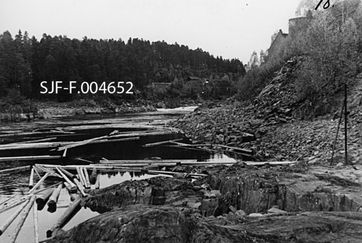 Drammenselva nedenfor Geithusfossen på Modum i Buskerud, fotografert ved sjeldent lav vannstand 21. oktober 1945.  Bildet er tatt fra noen fjellskjær som stikker ut i elveløpet på sørsida av en elvesving nedenfor Drammenselvens Papirfabrikker.  Vi skimter et par av bygningsgavlene på fabrikken på bakkekammen til høyre i bildeflata.  I forgrunnen ligger en del tømmer og flyter på et forholdsvis stilleflytende elveparti.  Lengre oppe skimter vi kvitskummende vann i Hårkallfossen, det nedre fallet i Geithusfossen.  Til venstre i bakgrunnen ser vi Hontvetåsen, hvor vi også skimter litt av traséen for Randsfjordbanen. 