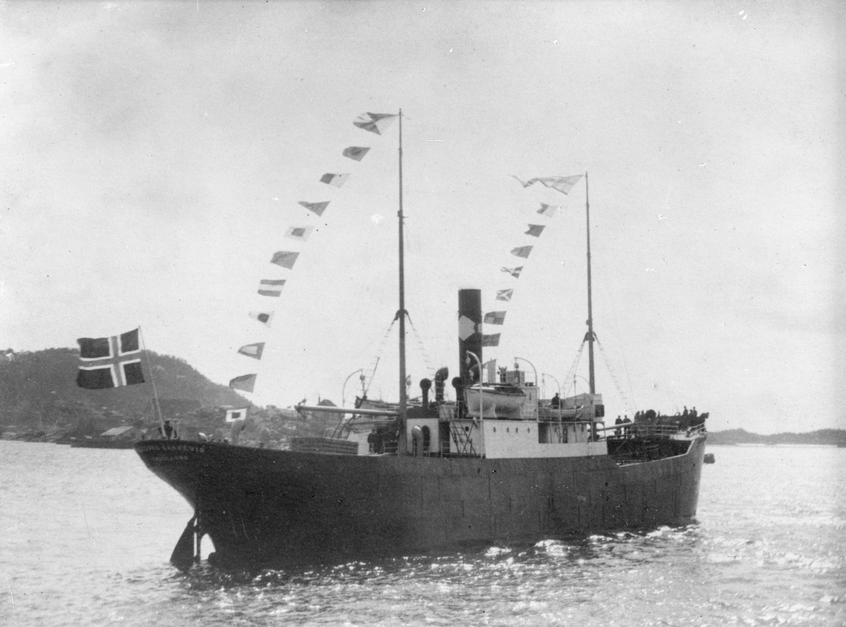 Dampskipet D/S "Ingeborg Bakkevig", med deler av mannskapet i baugen, på dekk og akter. Sjøhus og fjell i bakgrunnen.