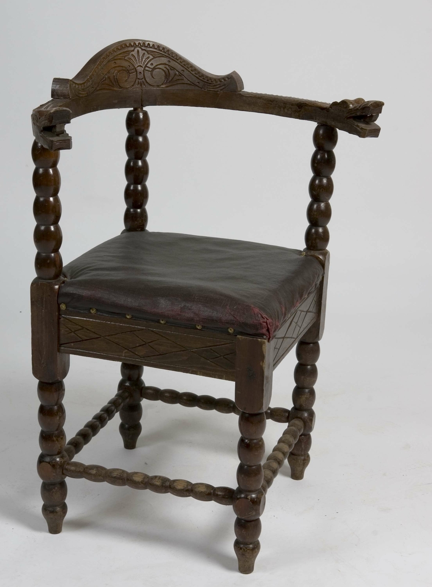 Trestol med diagonalt plassert, firkantet sete med mørkerød voksduk og blomsterformede nagler. Med kuleformer langs bein, sprosser, samt vertikal ryggdel. Øvre del av ryggen er halvsirkelformet og går over til armlenene som har et utskjært dragehode i hver ende.