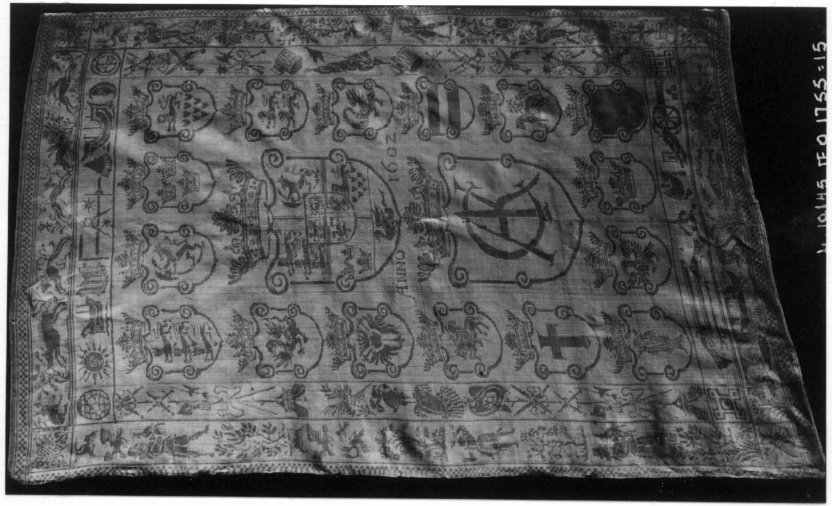 Det danske kongevåpen og Christian IV og Anna Catrines kronede navnetrakk omgitt av 16 skjold med alle de dansk-norske provinsers forskjellige våpenmerker. Dobbelt bord med våpentroféer, astronomiske instrumenter, jaktscener og fiskescener. Innvevd 1602 og brodert "H S II" i røde korssting.
